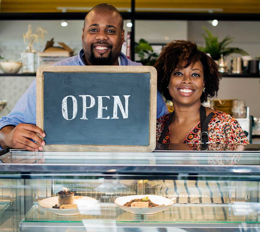 Man & wife holding a sign that says "Open" on top of a glass case with pastries